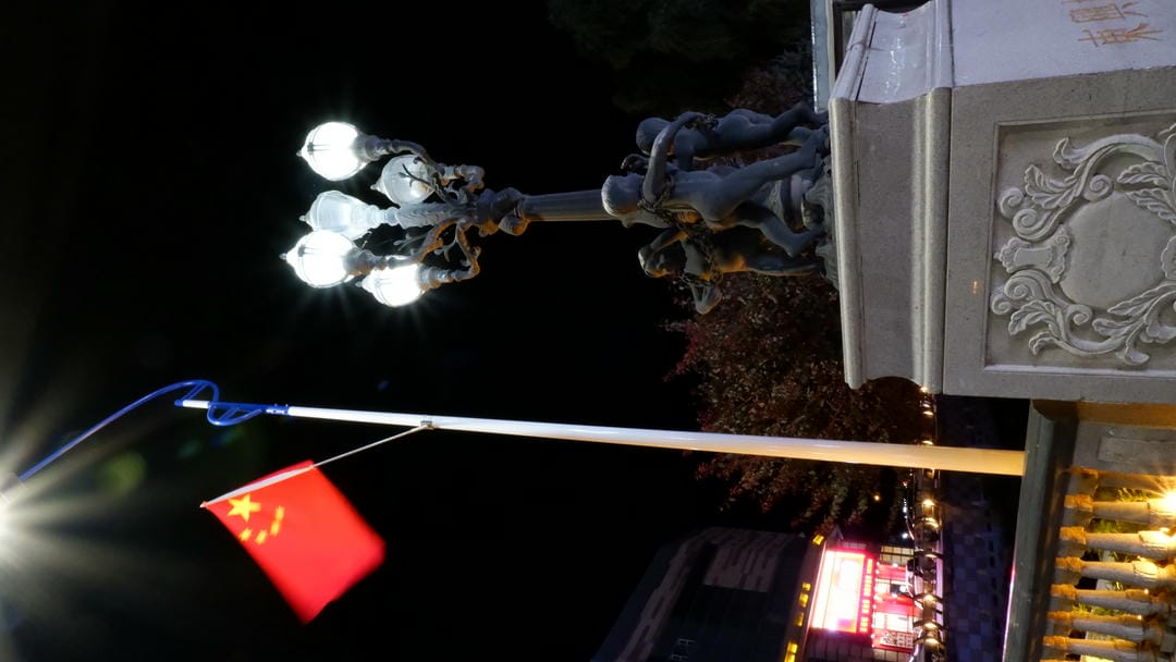 中国国旗在夜晚随风飘扬，旁边是装饰性的路灯。