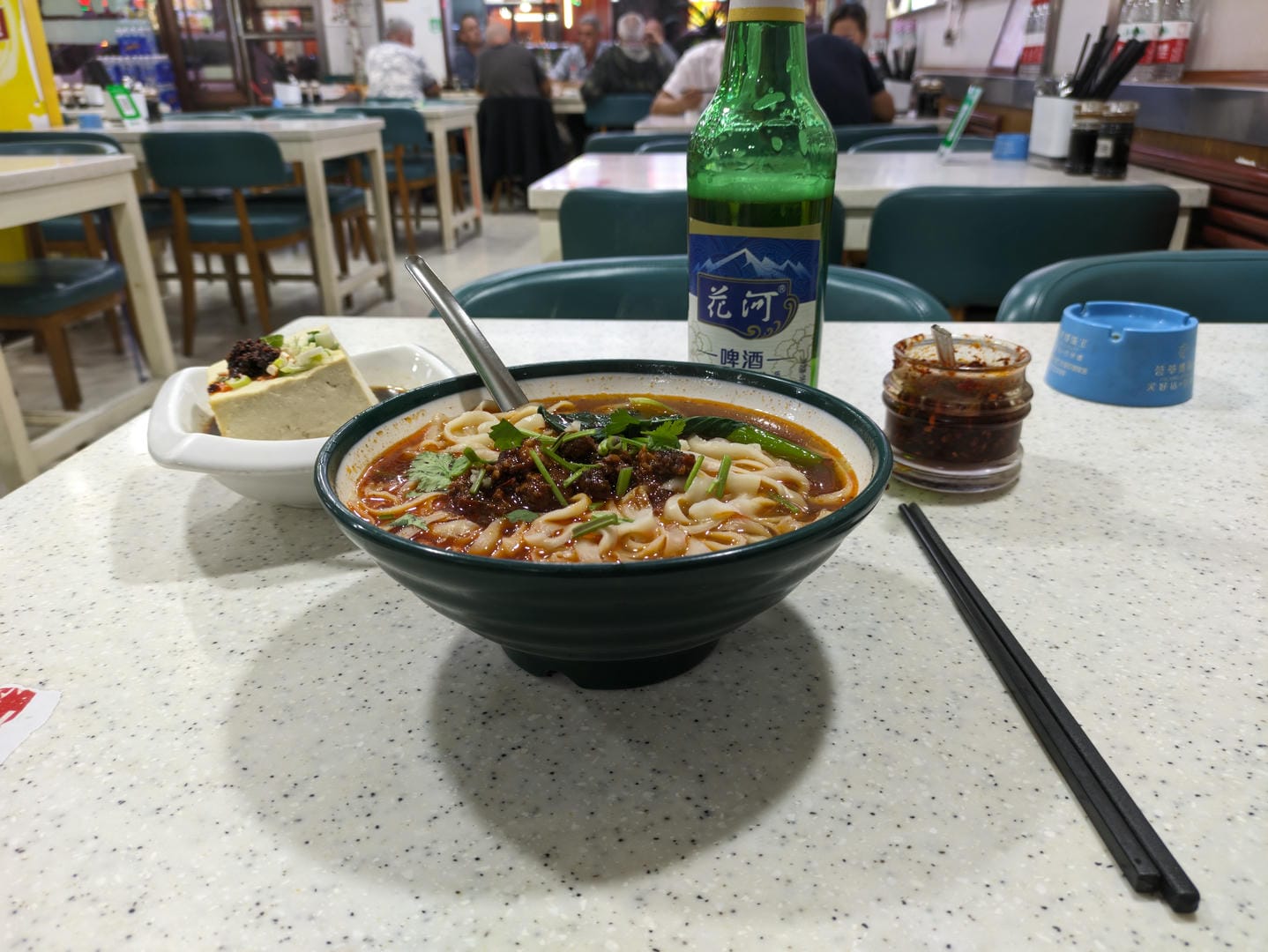 桌上有一碗面条、一瓶啤酒、一碟豆腐和辣椒酱。