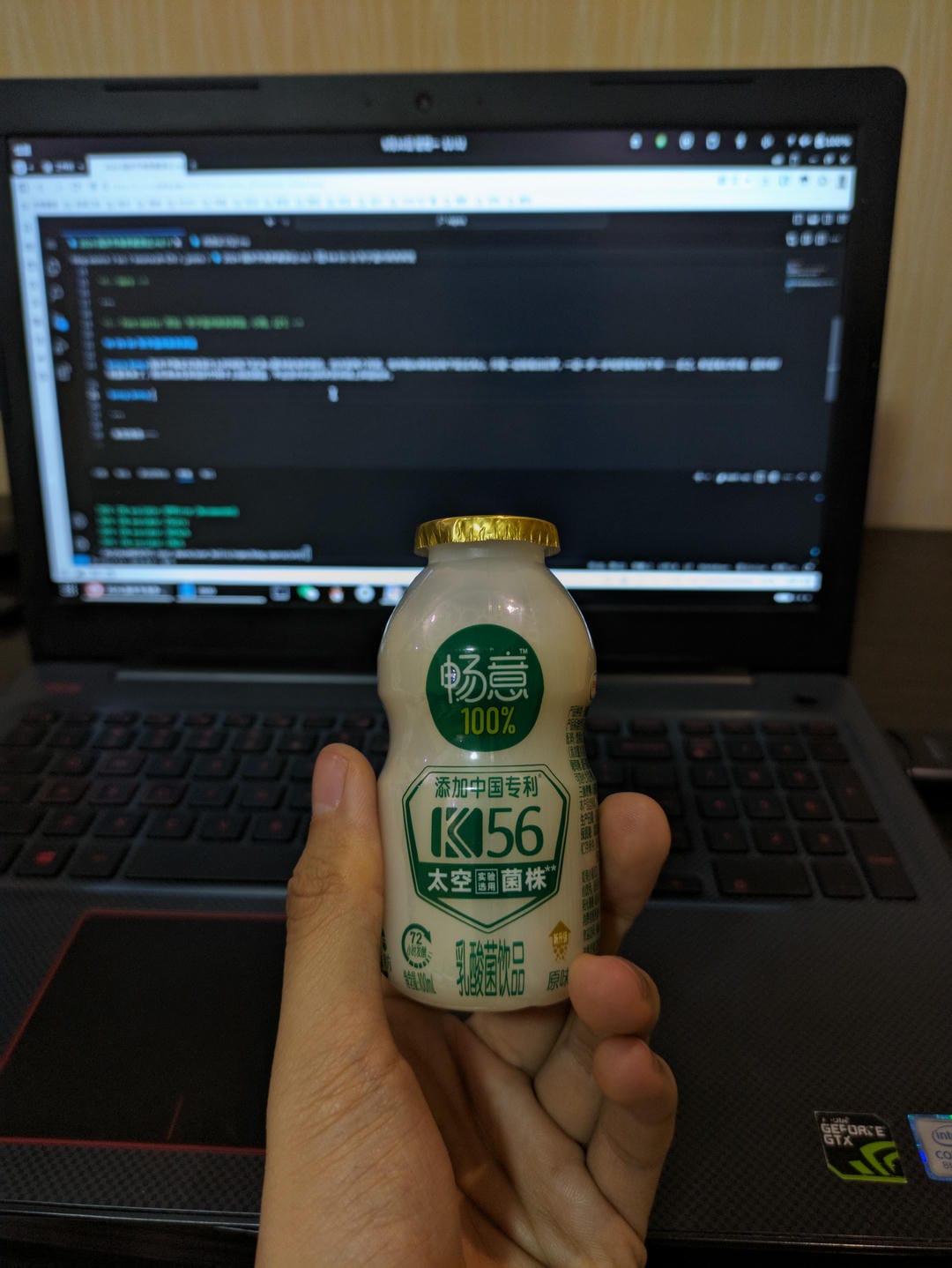 一只手拿着一瓶乳酸菌饮料,背景是一台笔记本电脑。