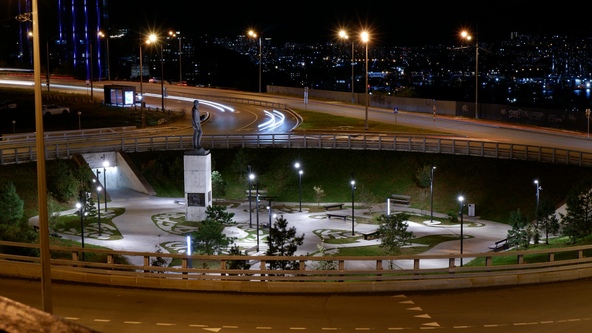 夜晚的城市公园，中央有一座雕像，周围有灯光照亮的小径和树木。