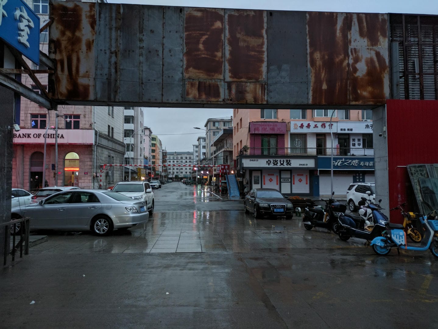 雨后的街道,地面湿滑,两侧停着汽车和摩托车,上方有生锈的金属结构。