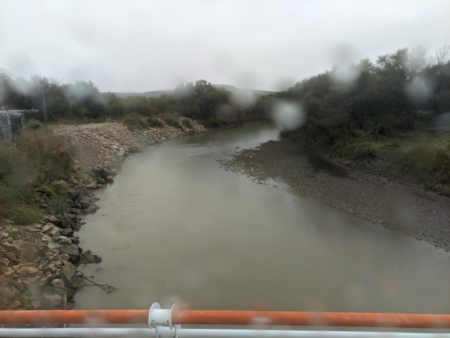 一条河流,河水浑浊,河岸两侧是岩石和树木,前景有栏杆。