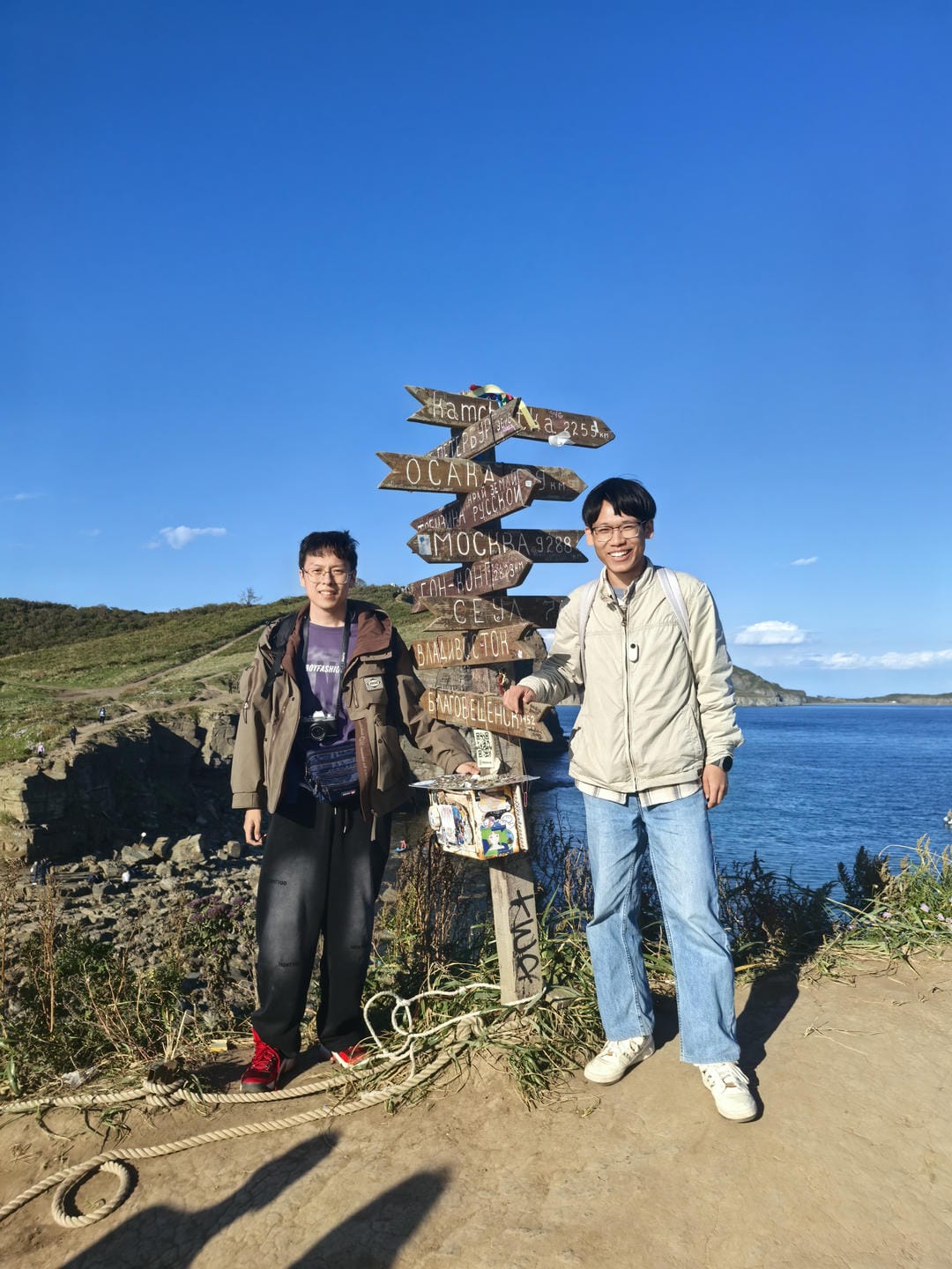 两个人站在海边,背景是蓝色的天空和海洋。他们身后有一个指示牌,上面写着不同城市的距离。左边的人穿着棕色外套和黑色裤子,右边的人穿着浅色夹克和蓝色牛仔裤。