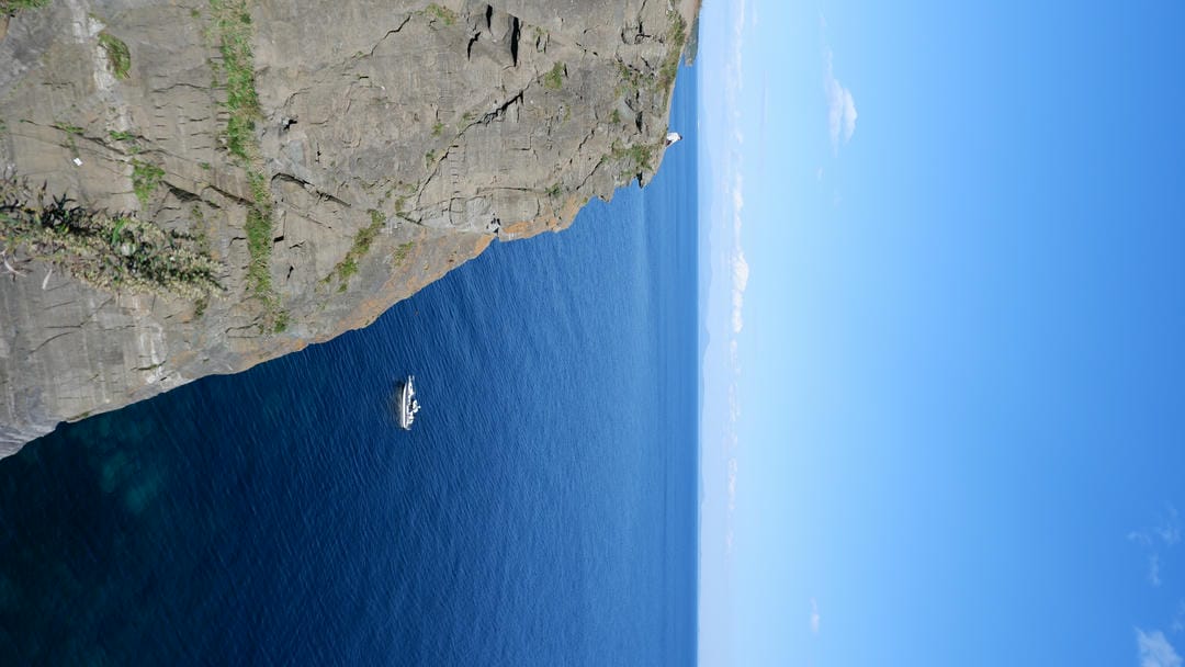 悬崖边有一艘小船停泊在深蓝色的海面上，远处可以看到山脉。