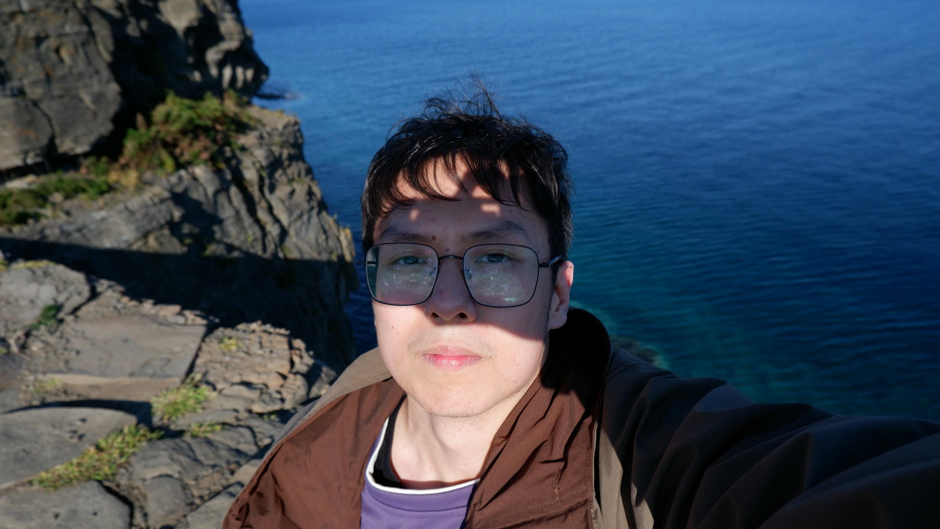 一个戴眼镜的人站在悬崖边，背景是深蓝色的大海和岩石。