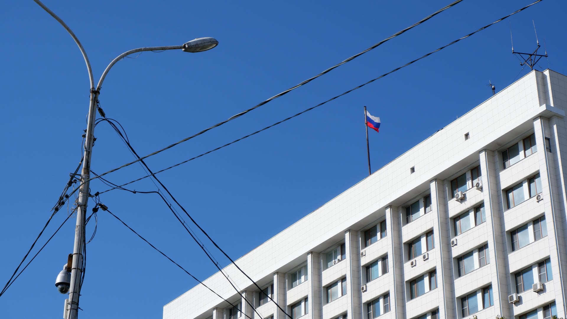 一面俄罗斯国旗飘扬在建筑物顶部，建筑物前有电线杆和路灯。