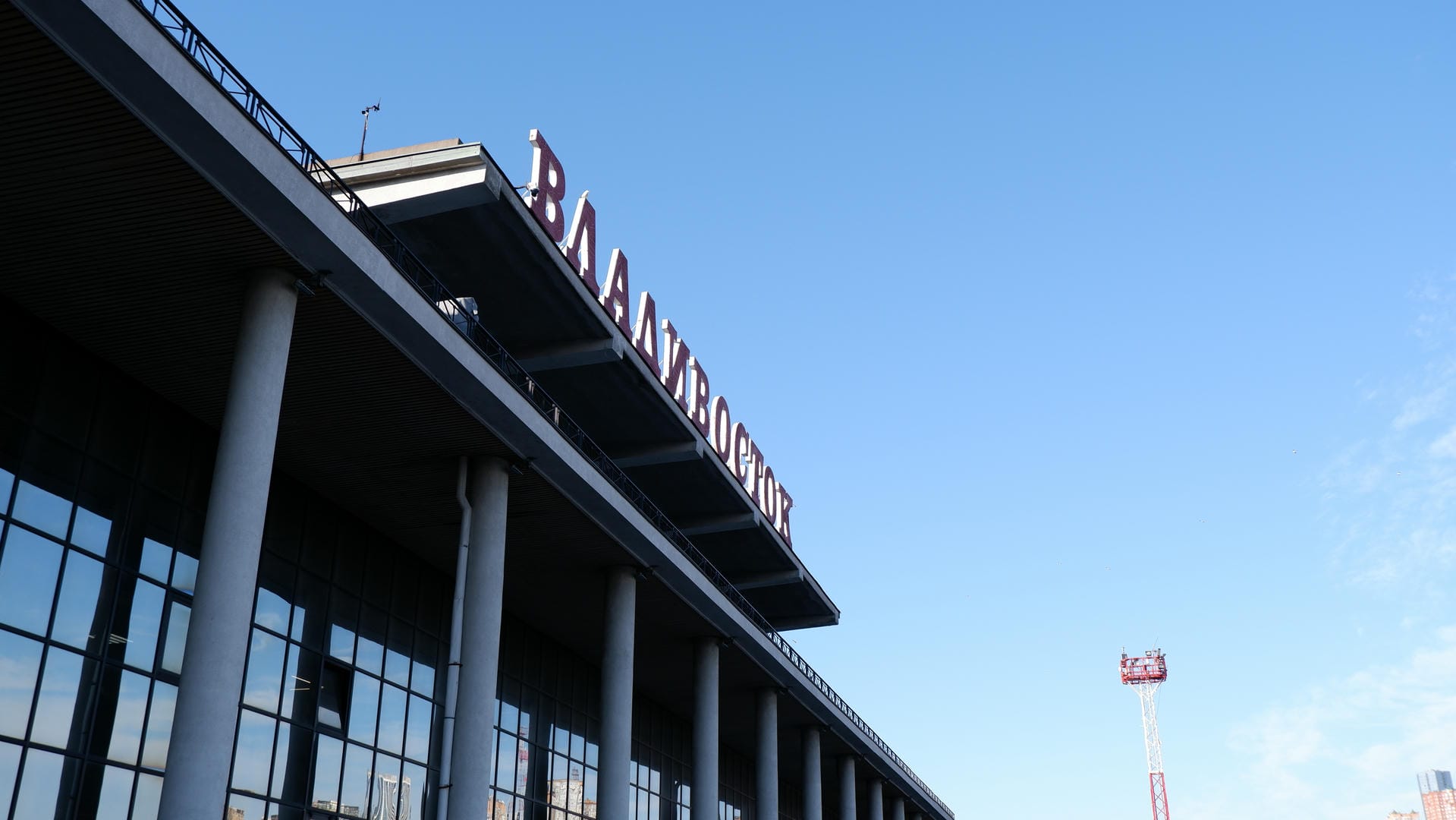 一座现代建筑的顶部，上面有大大的红色字母写着“ВЛАДИВОСТОК”，背景是蓝天。