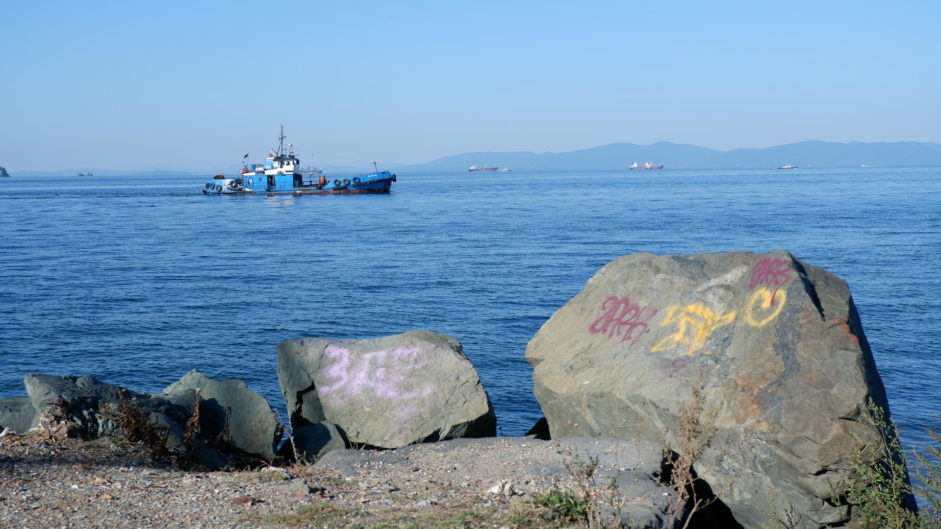 海岸边有几块大石头，上面涂有粉色和黄色的涂鸦，远处有一艘蓝色的船。
