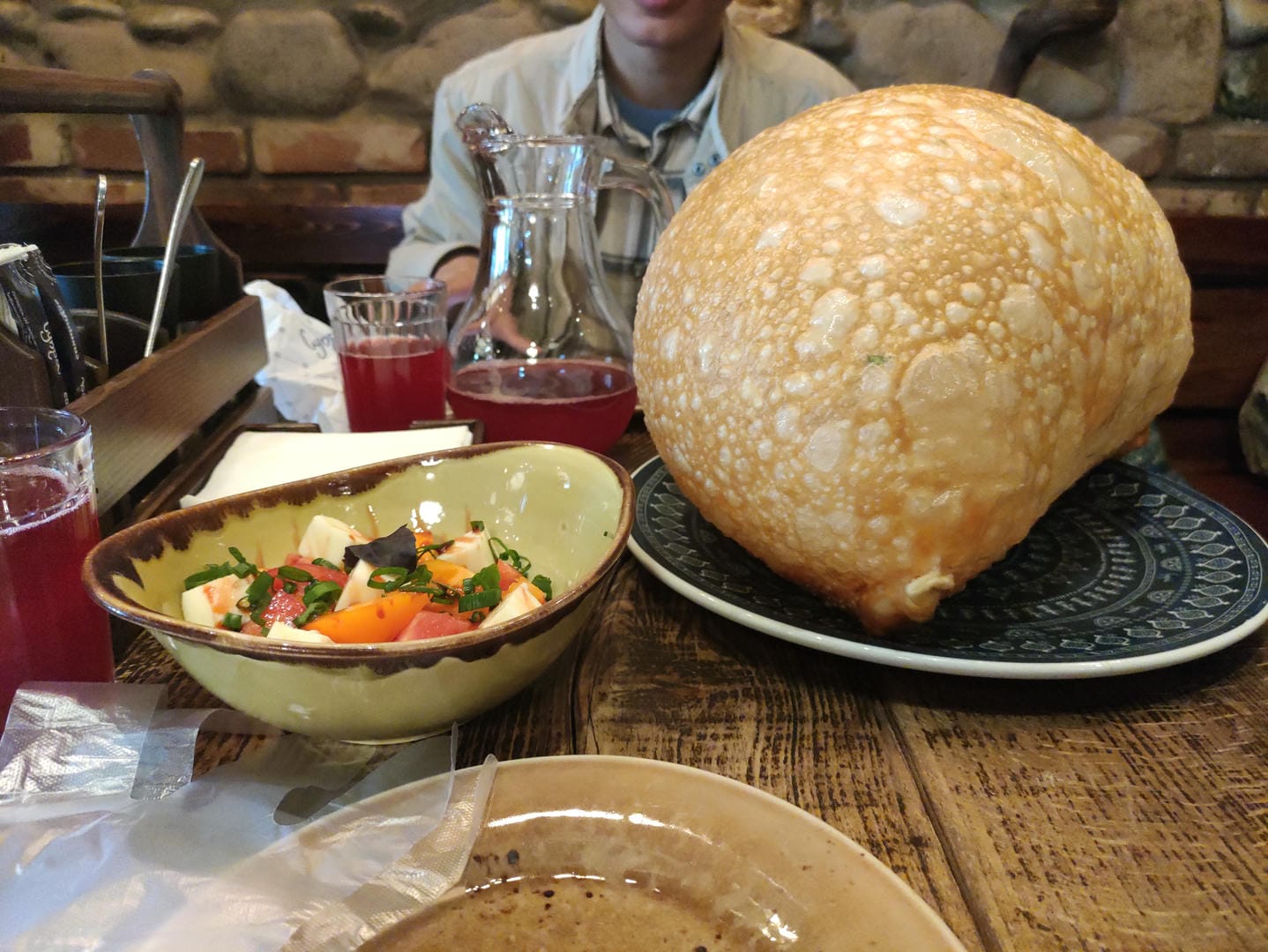 餐桌上有一个大面包、一碗沙拉和一杯红色饮料,背景中有人坐着。