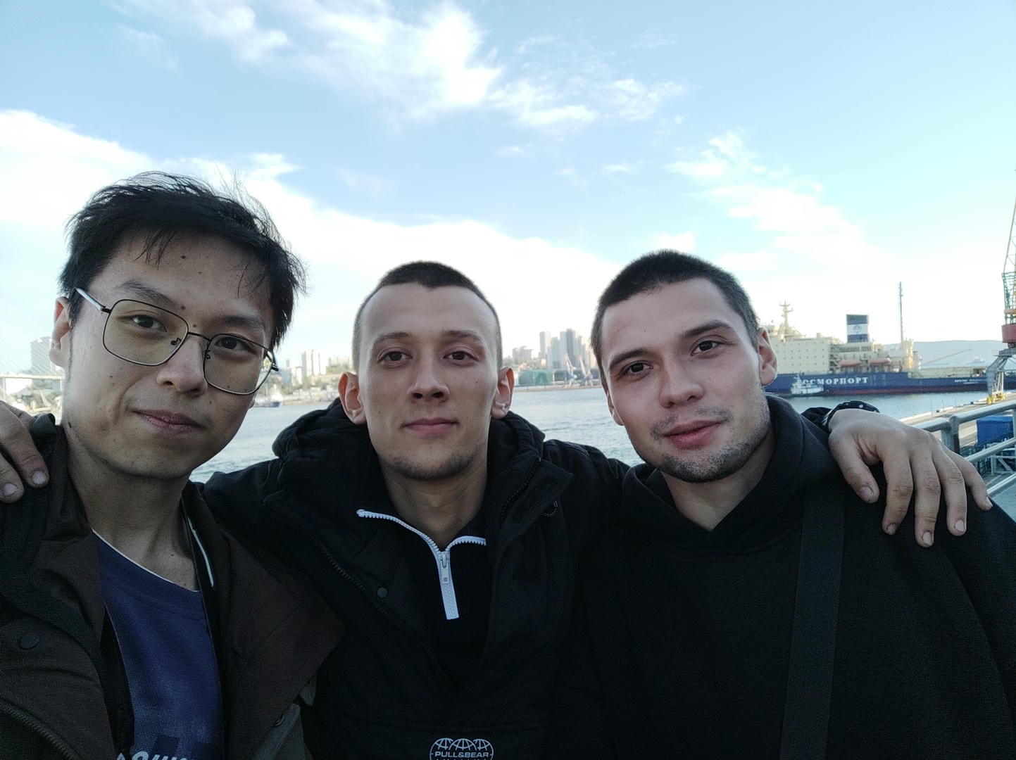三个人站在户外合影,背景是水面和远处的城市建筑。