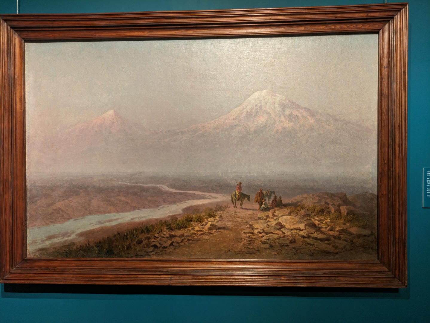 画框内展示了一幅描绘山景的油画,前景有几个人和马匹。
