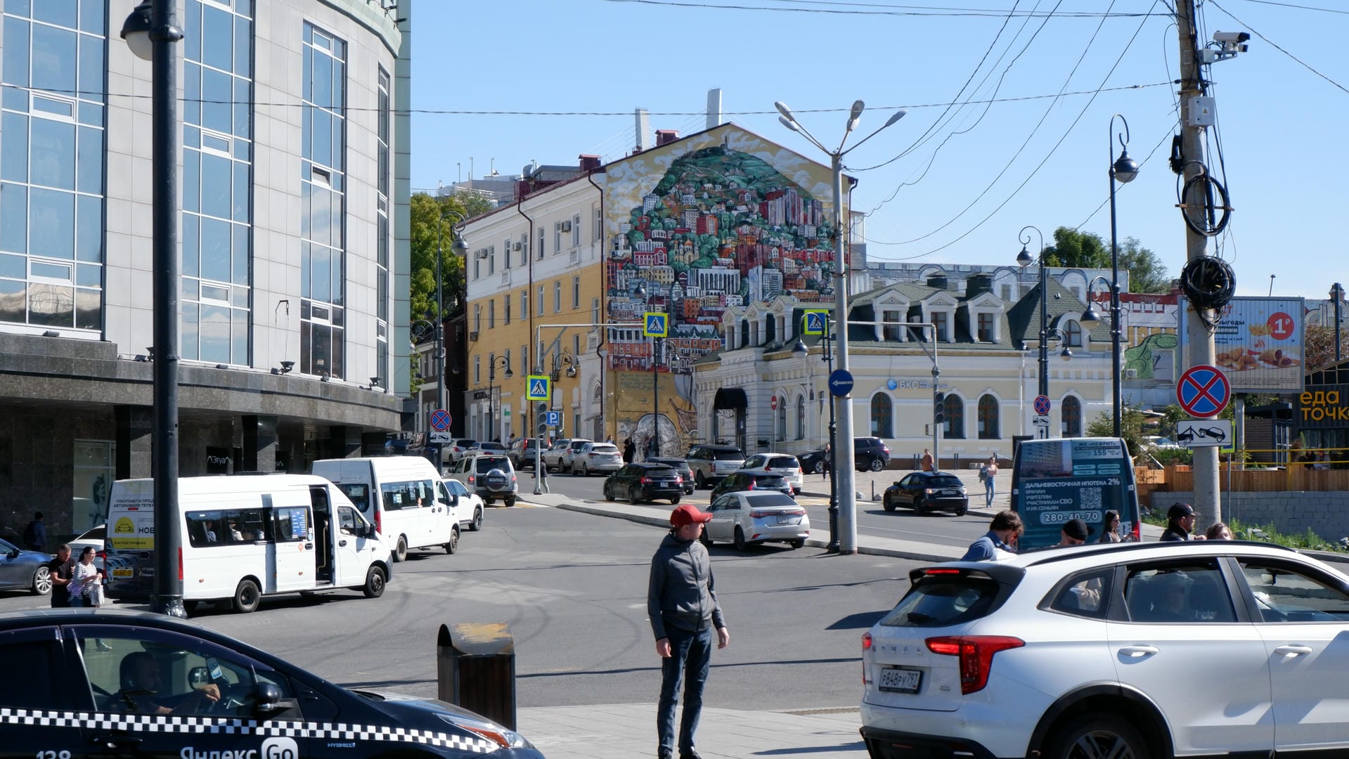 繁忙的城市街道，左侧是现代建筑，右侧有大型壁画装饰的旧建筑，街道上有行人和车辆。