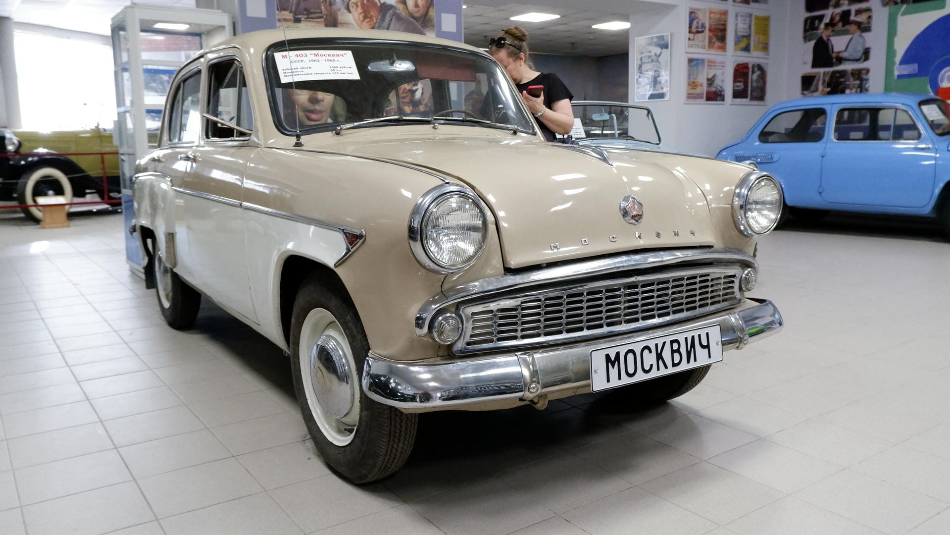 一辆米色的复古汽车，车牌上写着“МОСКВИЧ”，停放在室内展览空间。