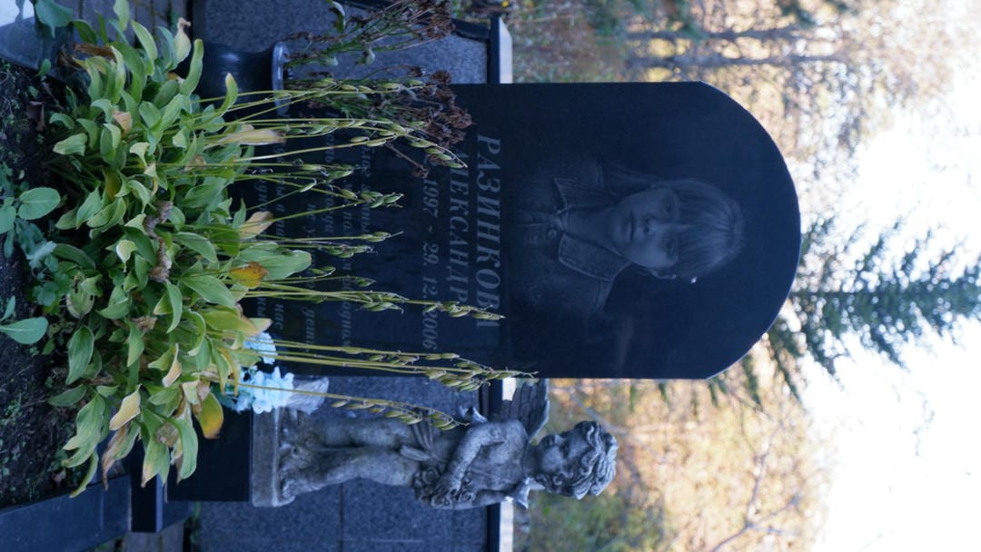 一块黑色墓碑上刻有小女孩的照片和名字“Разинкова”，旁边有一个小天使雕像。