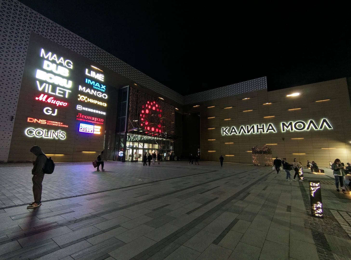 夜晚的城市广场上有许多商店招牌亮着灯,人们在广场上行走。