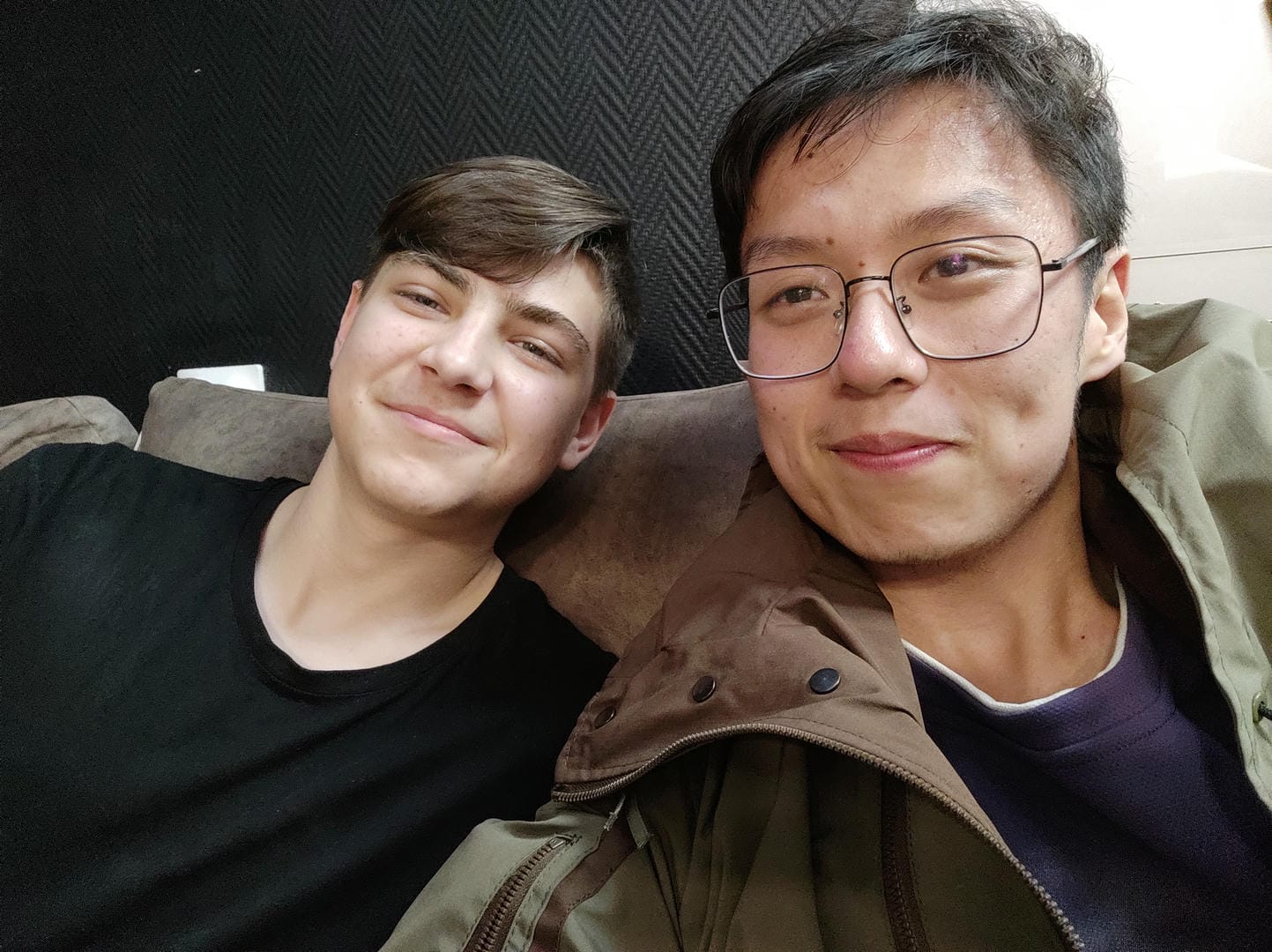 两个人坐在沙发上,左边的人穿着黑色上衣,右边的人戴着眼镜,穿着棕色外套。