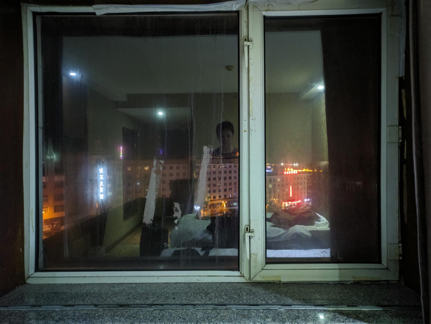 透过窗户看到房间内有人躺在床上,窗外是城市的夜景。