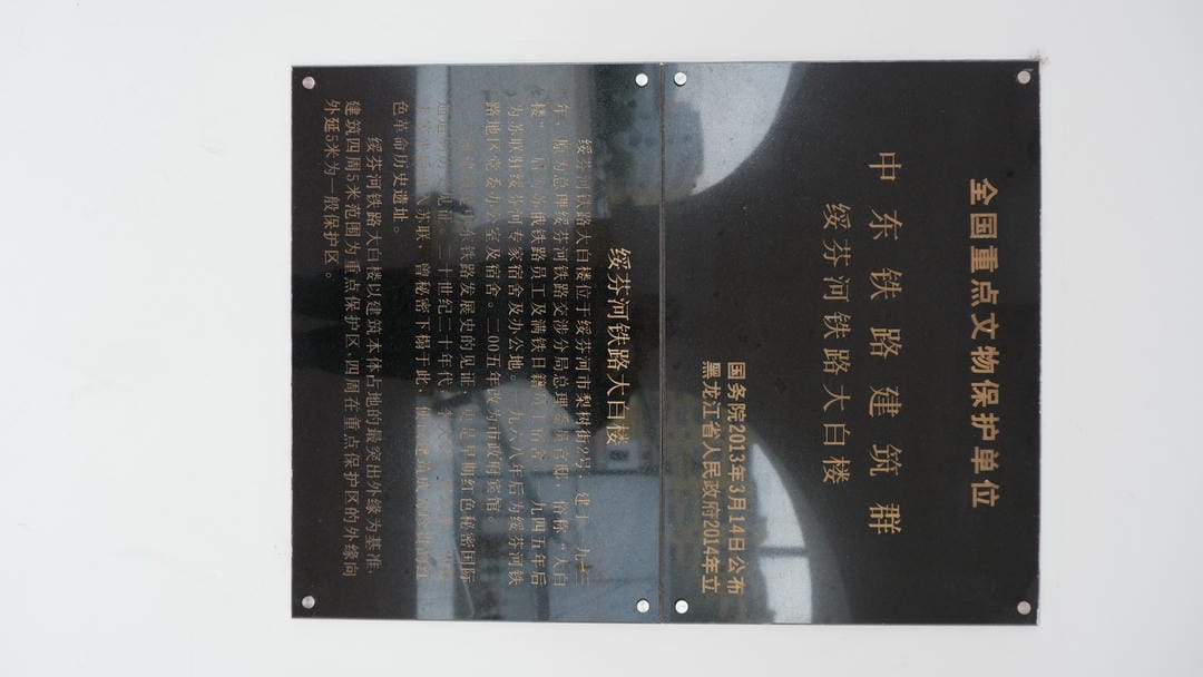 一块黑色金属牌匾，上面用中文写着“全国重点文物保护单位”以及“中东铁路建筑群 绥芬河铁路大白楼”等信息。
