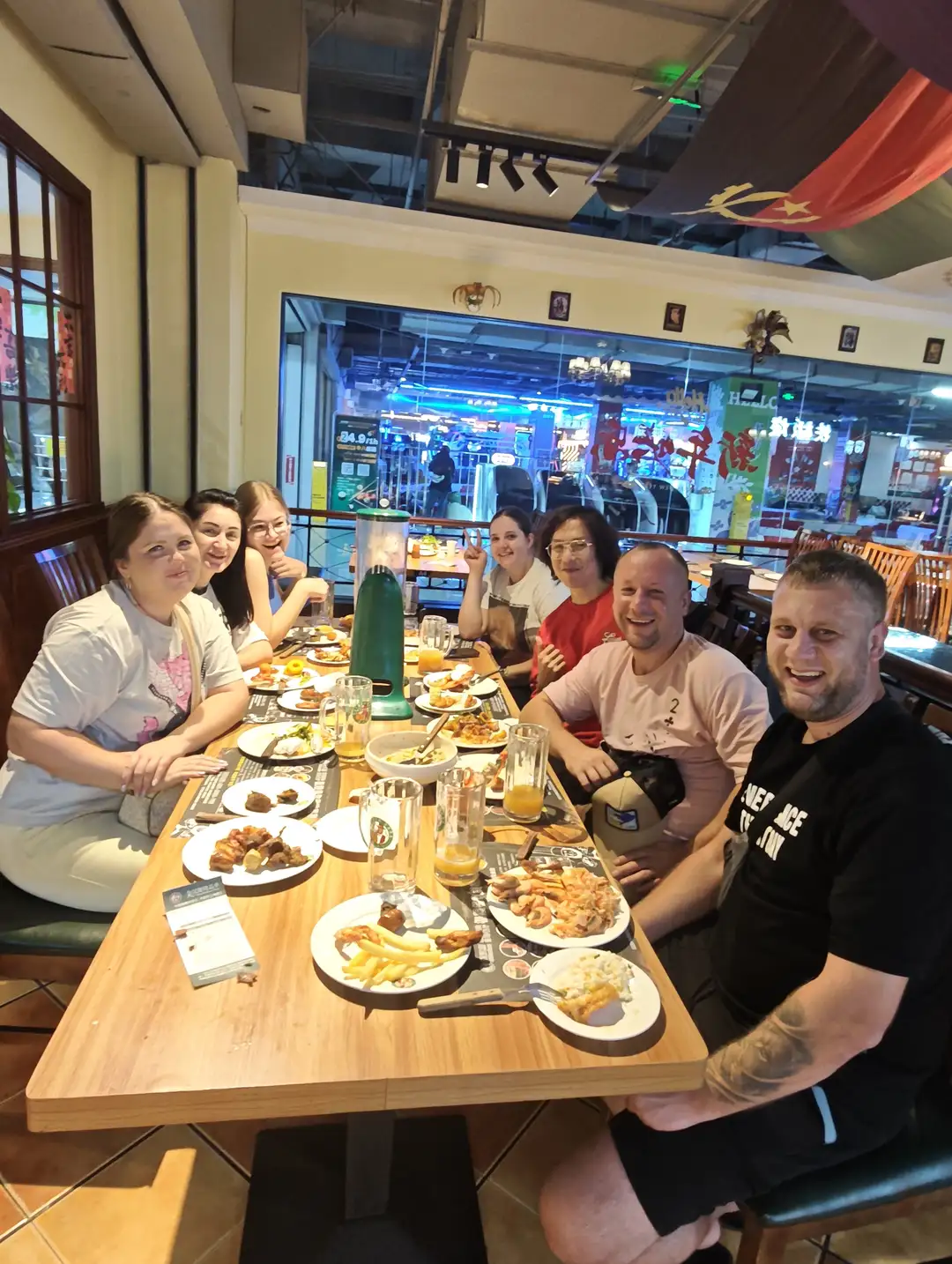 A group of people are sitting around a long table inside a restaurant, with various dishes and drinks laid out on the table, including french fries, meat, and orange juice. They are dressed casually and have cheerful expressions; some are making victory gestures, while others are smiling at the camera. The restaurant environment is spacious and bright, with red flags hanging from the ceiling, and other dining areas and shops visible in the background.