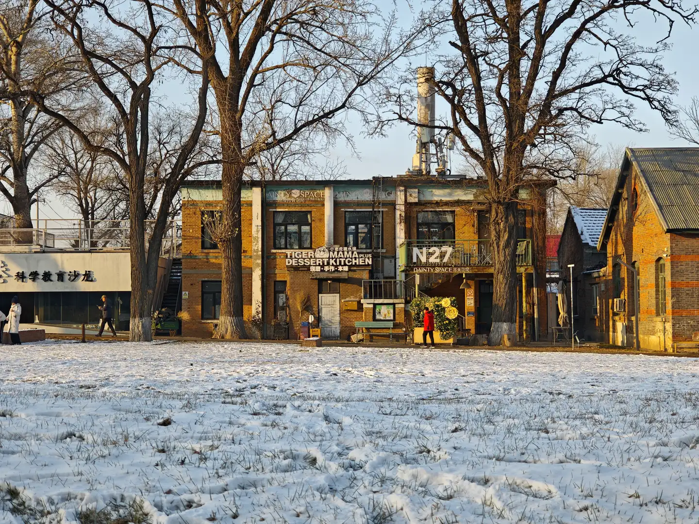 Snow-dusted grassy area in front of a vibrant brick building housing cafes. Tall, bare trees, people walking, and a clear blue sky create a cozy winter scene.